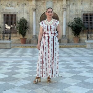 Mujer posando de cuerpo entero con vestido largo blanco estampado floral burdeos y sandalias doradas en plaza adoquinada