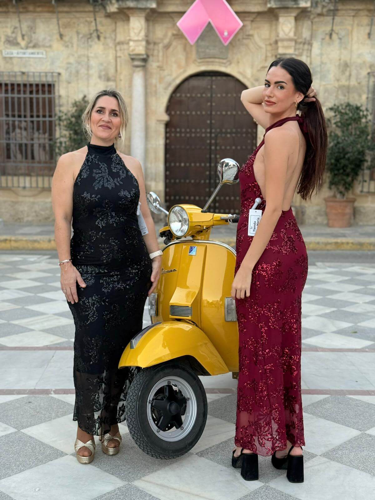 Dos mujeres con vestido largo burdeos y negro de lentejuelas y cuello halter, fondo urbano con moto amarilla